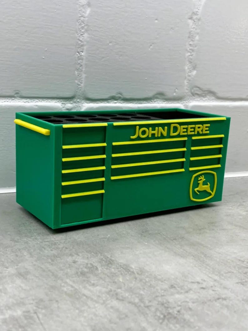 Może przedstawiać: Zielone i ż&oacute;łte prostokątne pudełko do przechowywania z napisem "JOHN DEERE" i logo. Pudełko ma ż&oacute;łte poziome paski i ż&oacute;łty uchwyt. Pudełko stoi na szarej powierzchni.