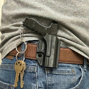 Könnte beinhalten: Schwarzer, 3D-gedruckter, pistolenförmiger Schlüsselanhänger, der an einer Gürtelholster befestigt ist. Die Holster ist an einem braunen Ledergürtel befestigt, der mit einer blauen Jeans getragen wird. Ein Satz Schlüssel hängt am Schlüsselanhänger.
