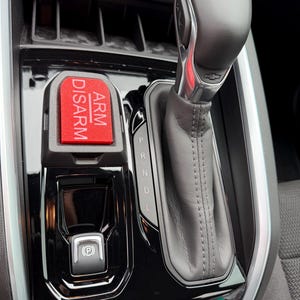 May include: Interior car console featuring a gear shift lever, a red button with "ARM DISARM" text, and a parking brake button. The console is black with silver accents and a Chevrolet logo.