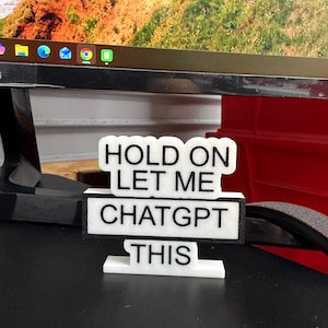 May include: A white and black desk sign with the text "HOLD ON LET ME CHATGPT THIS". The sign is constructed from layered materials and is positioned on a desk. A computer monitor and a red object are visible in the background.