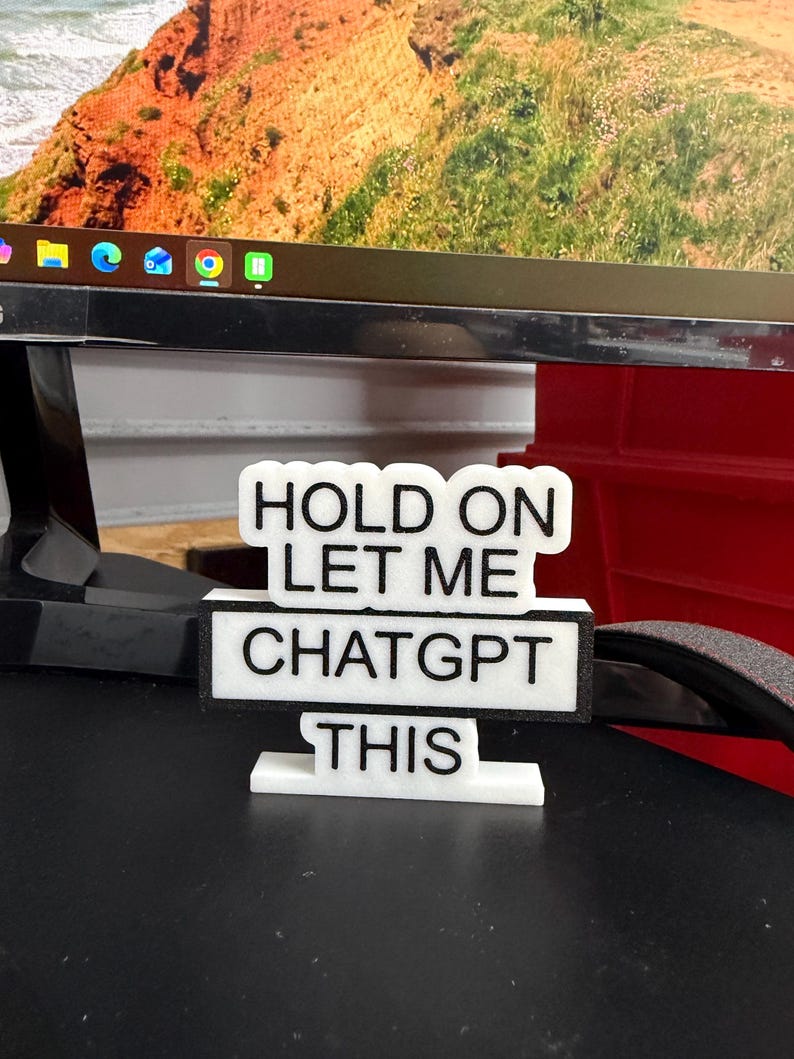 May include: A white and black desk sign with the text "HOLD ON LET ME CHATGPT THIS". The sign is constructed from layered materials and is positioned on a desk. A computer monitor and a red object are visible in the background.