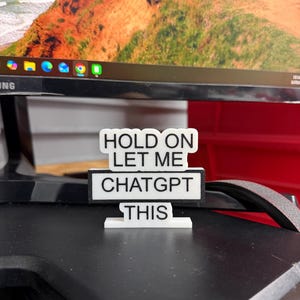 May include: A white and black sign with the text "HOLD ON LET ME CHATGPT THIS". The sign is on a black surface, with a computer monitor in the background.