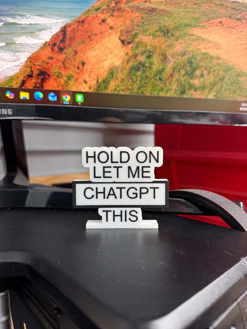 May include: A white and black sign with the text "HOLD ON LET ME CHATGPT THIS". The sign is on a black surface, with a computer monitor in the background.