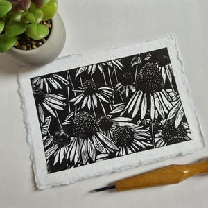 Puede incluir: Un grabado en linóleo en blanco y negro de flores de equinácea sobre papel texturizado. La obra de arte presenta un diseño floral detallado con un fondo oscuro y pétalos blancos. También se ve una herramienta de tallado de madera y una pequeña planta suculenta en maceta.