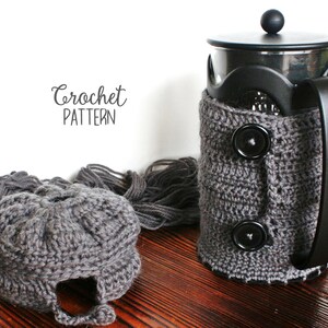 May include: A gray crocheted cozy for a French press coffee maker. The cozy has two black buttons and a crocheted border. The text "Crochet Pattern" is written on a white background.