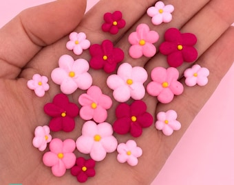 以下が含まれることがあります： ピンクやマゼンタの様々な色合いをした、5枚の花びらを持つ小さな花のデコレーションのコレクション。それぞれに小さな黄色い中心部があります。これらの装飾品はグループに配置されており、異なるサイズと鮮やかな色調を示しています。