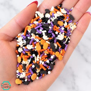 May include: A collection of Halloween-themed sprinkles featuring orange pumpkin shapes, white ghost figures, and black, purple, and orange rod-shaped jimmies mixed with small white nonpareils. The mix includes various textures and sizes of sugar candy.