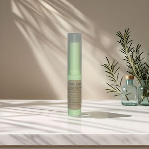 Può includere: Un tubo verde di balsamo labbra vegano con il testo "Vegan Lip Balm" e "Enriched with Chamomile, Glycerin & Vitamin E" sull'etichetta. Il tubo è su una superficie bianca con un rametto di rosmarino sullo sfondo.