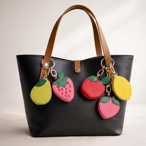 May include: Black tote bag with brown handles and gold hardware, adorned with fruit-shaped keychains. The keychains include a yellow pear, a pink strawberry, a red apple, a pink peach, and a yellow lemon.
