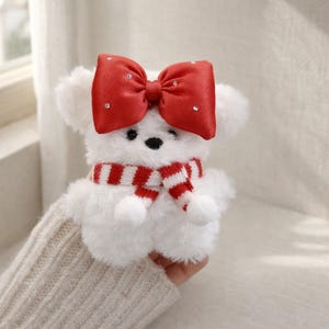 May include: A white plush bear with a red bow adorned with rhinestones and a red and white striped scarf. The bear has black eyes and a black nose. The bear is a cute and cuddly toy.