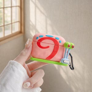May include: Pink AirPod case shaped like a cartoon snail, featuring a red swirl design and blue accents. The case has green eyes and a green base, with a black carabiner clip attached.