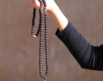 Puede incluir: Un collar largo de cuentas de madera marrón oscuro con cuentas de acento rojas y turquesas. El collar está sostenido por una mano, y el fondo es marrón borroso.