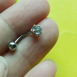 Pode incluir: Uma joia corporal de barra curva feita de metal prateado polido. Uma extremidade apresenta uma esfera lisa e redonda, enquanto a outra extremidade exibe uma pequena pedra cabochão azul-clara translúcida presa por quatro garras de metal.