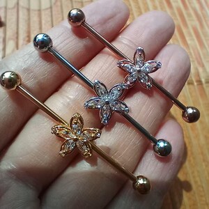 May include: Three industrial barbells in gold, silver, and rose gold. Each barbell has a small ball on each end and a flower-shaped charm in the centre. The flower charms are adorned with clear, sparkling stones.
