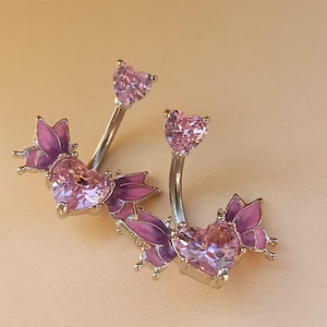 May include: Two belly button rings featuring heart-shaped pink gemstones and butterfly designs. The butterflies have pink and purple wings. The metal is silver-coloured.