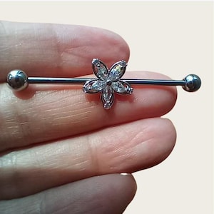 May include: A silver industrial barbell earring with a flower design and clear, sparkling accents. The barbell has a ball on each end. The flower is the focal point, adding a decorative touch to the body jewellery.