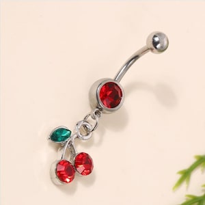 Puede incluir: Anillo de ombligo plateado con un colgante de dos cerezas rojas con hojas verdes y brillantes transparentes.