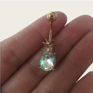 Könnte beinhalten: Goldener Bauchnabelring mit einem klaren, schillernden Ananas-Anhänger. Der Ananas-Anhänger hat eine goldene Spitze und einen klaren Stein in der Mitte.