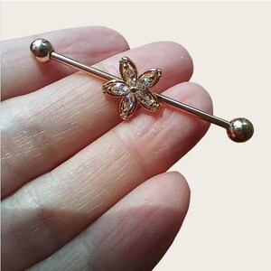 May include: A gold-coloured industrial barbell earring. The barbell features a flower-shaped charm with clear, sparkling stones at the centre. The barbell has a ball on each end.