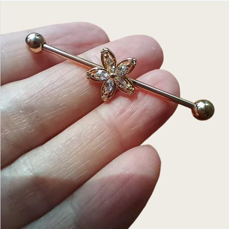 May include: A gold-coloured industrial barbell earring. The barbell features a flower-shaped charm with clear, sparkling stones at the centre. The barbell has a ball on each end.