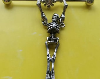 May include: Silver industrial barbell featuring a skeleton charm. The barbell has a small, clear gem in the center and round ball ends. The skeleton is detailed with visible ribs and leg bones. The jewelry is set against a yellow background.