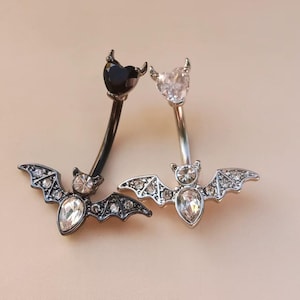 Puede incluir: Dos piercings para el ombligo con diseños de murciélagos. Un anillo es negro con una gema negra en forma de corazón y un murciélago con gemas transparentes. El otro anillo es plateado con una gema transparente en forma de corazón y un murciélago con gemas transparentes.