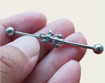 May include: A silver industrial barbell earring featuring a detailed lizard charm in the center. The barbell has a straight bar with a ball on each end. The lizard is a detailed, three-dimensional design.