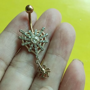 May include: A gold-toned navel ring featuring a heart-shaped spider web design encrusted with clear rhinestones. A small heart-shaped stone sits at the center of the web, and a dangling gold-toned spider charm hangs from the bottom of the web.