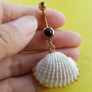 May include: A gold-toned navel ring featuring a curved barbell with a small gold ball at the top and a larger black circular stone in the centre. A white seashell with a gold-toned metallic rim hangs from the bottom of the barbell.