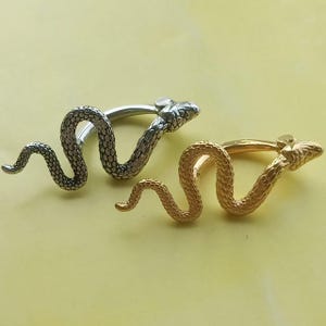 May include: Two snake-shaped rings. One ring is silver-colored, and the other is gold-colored. The snakes are coiled with detailed scales and have a curved design. The background is a solid light yellow.