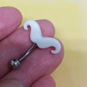 Op de afbeelding: Een gebogen wenkbrauwring met een wit snorontwerp. De sieraden hebben een zilverkleurige, gebogen halter met een kleine, ronde bal aan één uiteinde. De snor is effen, glanzend wit.