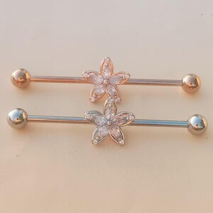 May include: Two industrial barbells. One is rose gold with a clear flower design, and the other is silver with a clear flower design. Each barbell has a ball on each end.