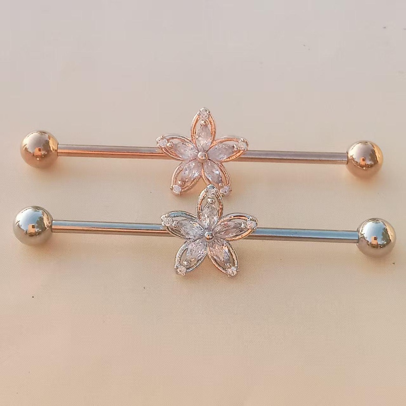 May include: Two industrial barbells. One is rose gold with a clear flower design, and the other is silver with a clear flower design. Each barbell has a ball on each end.