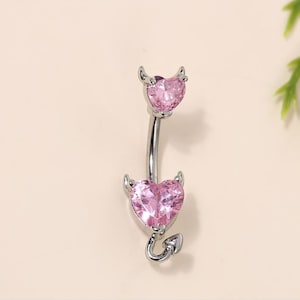 Può includere: Un piercing per l'ombelico argentato con due ciondoli a forma di cuore rosa. Ogni ciondolo ha piccole corna di diavolo.