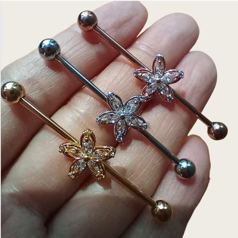 May include: Industrial barbell piercings in gold, silver, and rose gold. Each barbell features a flower design with clear crystals. The bars have a ball on each end. The jewellery is shown against a neutral background.