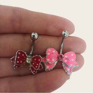 Puede incluir: Dos anillos de ombligo con diseño de lazo. Un lazo es rosa con lunares blancos y el otro es rojo con lunares blancos. Ambos lazos tienen un strass transparente en el centro.