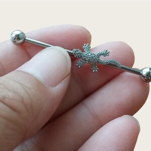May include: A silver industrial barbell earring featuring a lizard-shaped charm. The charm is encrusted with small, sparkling stones. The barbell has a smooth, polished finish with a ball on each end.