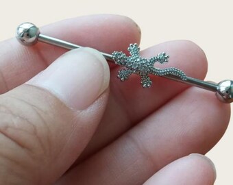 May include: A silver industrial barbell earring featuring a lizard-shaped charm. The charm is encrusted with small, sparkling stones. The barbell has a smooth, polished finish with a ball on each end.