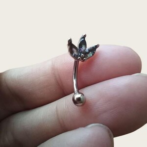 Puede incluir: Piercing de ceja curvado con una barra curva plateada. La parte superior presenta un grupo de tres gemas oscuras, iridiscentes, en forma de pétalo. La parte inferior de la barra tiene una pequeña bola plateada redonda.