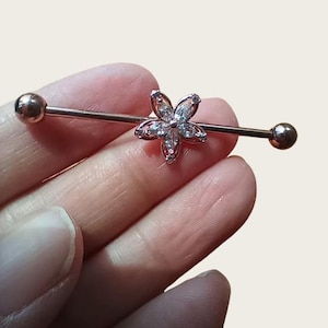 May include: A rose gold industrial barbell earring featuring a flower-shaped charm. The flower is embellished with clear, sparkling crystals. The barbell has a ball at each end. The jewellery is designed for body piercing.
