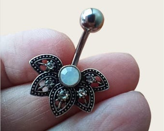 May include: A silver-colored belly button ring with a flower-shaped design. The flower has five petals with a filigree pattern and small clear gemstones. A larger, light-colored gemstone is in the center, and a silver ball tops the curved barbell.