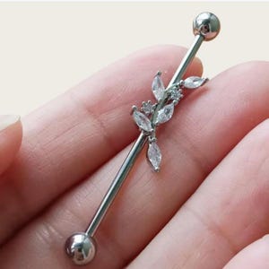 May include: A silver industrial barbell earring with a floral design. The barbell has a straight bar with a ball on each end. The floral design features clear, marquise-cut crystals arranged in a leaf pattern.