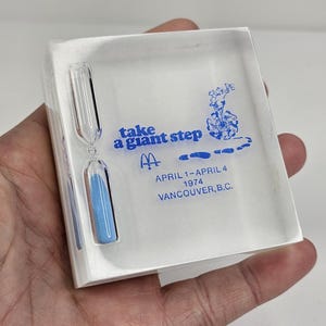 May include: Clear acrylic hourglass with blue sand, set within a clear rectangular block. The block features blue text that reads "take a giant step" and "April 1-April 4 1974 Vancouver, B.C.", along with a McDonald's logo.