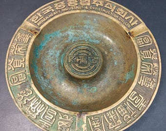 May include: Vintage brass ashtray with a circular design. The ashtray features embossed text in both English and Korean, with the text "KOREA HEAVY INDUSTRIES & CONSTRUCTION CO., LTD." and "KHC" in the center. The ashtray has a weathered, aged appearance.