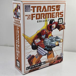 Peut inclure: Une boîte en carton rectangulaire pour The Transformers Collection 13, représentant un robot rouge et orange tenant un pistolet à double canon. L'emballage comporte le texte "More Than Meets The Eye" et des caractères japonais sur le devant.