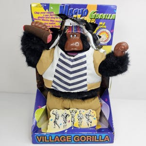 May include: A vintage "Macho Gorilla" toy in its original box. The plush gorilla wears a vest, shorts, and a hat. It holds a brown object in one hand and has a raised fist. The box features text and illustrations, including the words "Village Gorilla".