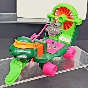 May include: A green and orange Teenage Mutant Ninja Turtles toy vehicle. The vehicle has pink wheels, a green foot-shaped front, and a green head with an orange mask. The vehicle has a silver engine and a pink and purple platform.