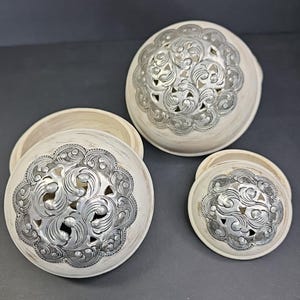 May include: A set of three cream-coloured decorative bowls with silver filigree designs. The bowls are round with a distressed finish, and the silver accents feature intricate floral patterns. One bowl has its lid open.