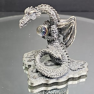 May include: A detailed silver dragon figurine with a multi-colored crystal orb. The dragon is posed on a textured base, with its wings spread and head raised. The sculpture has intricate scale and wing details.