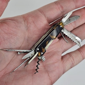 May include: A vintage multi-tool with a tortoiseshell handle. The tool features various blades, a corkscrew, and other implements. The metal components show signs of age and use. The tool is open, displaying its many functions.